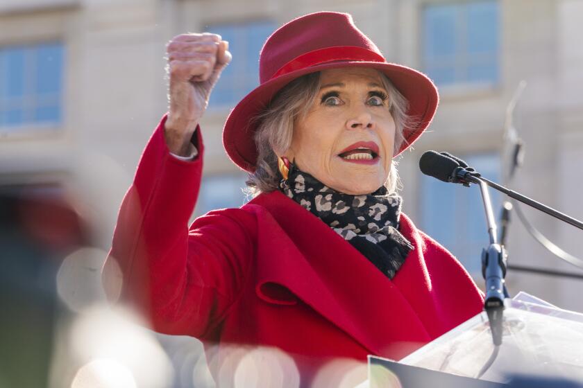 An older woman in a red coat and red hat speaks into a microphone and raises her right fist at a protest