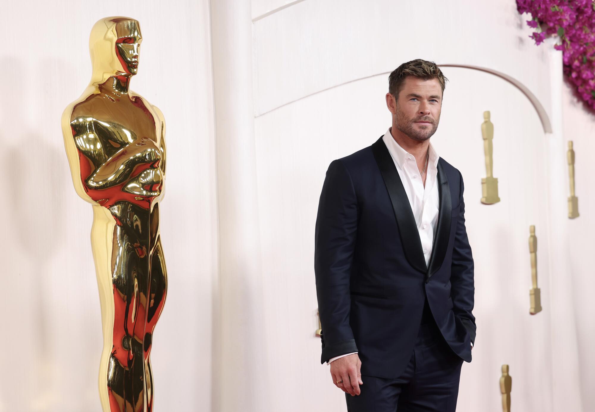 Chris Hemsworth wears a suit as he stands in front of an Oscar statue. 