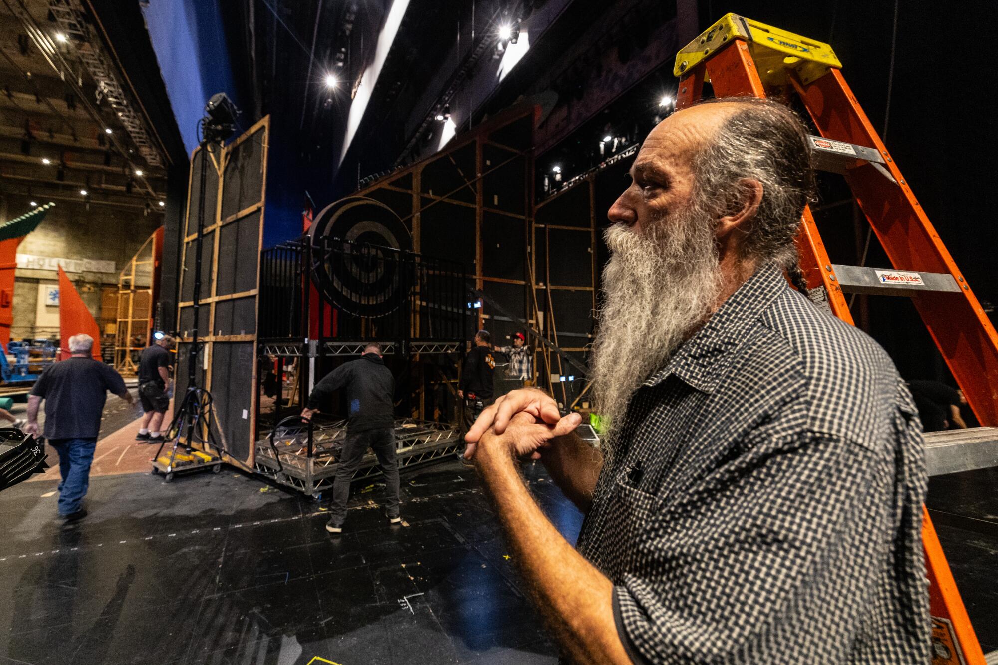 A man with flowing gray beard stands by a ladder and watches set pieces move around the Dorothy Chandler Pavilion stage. 