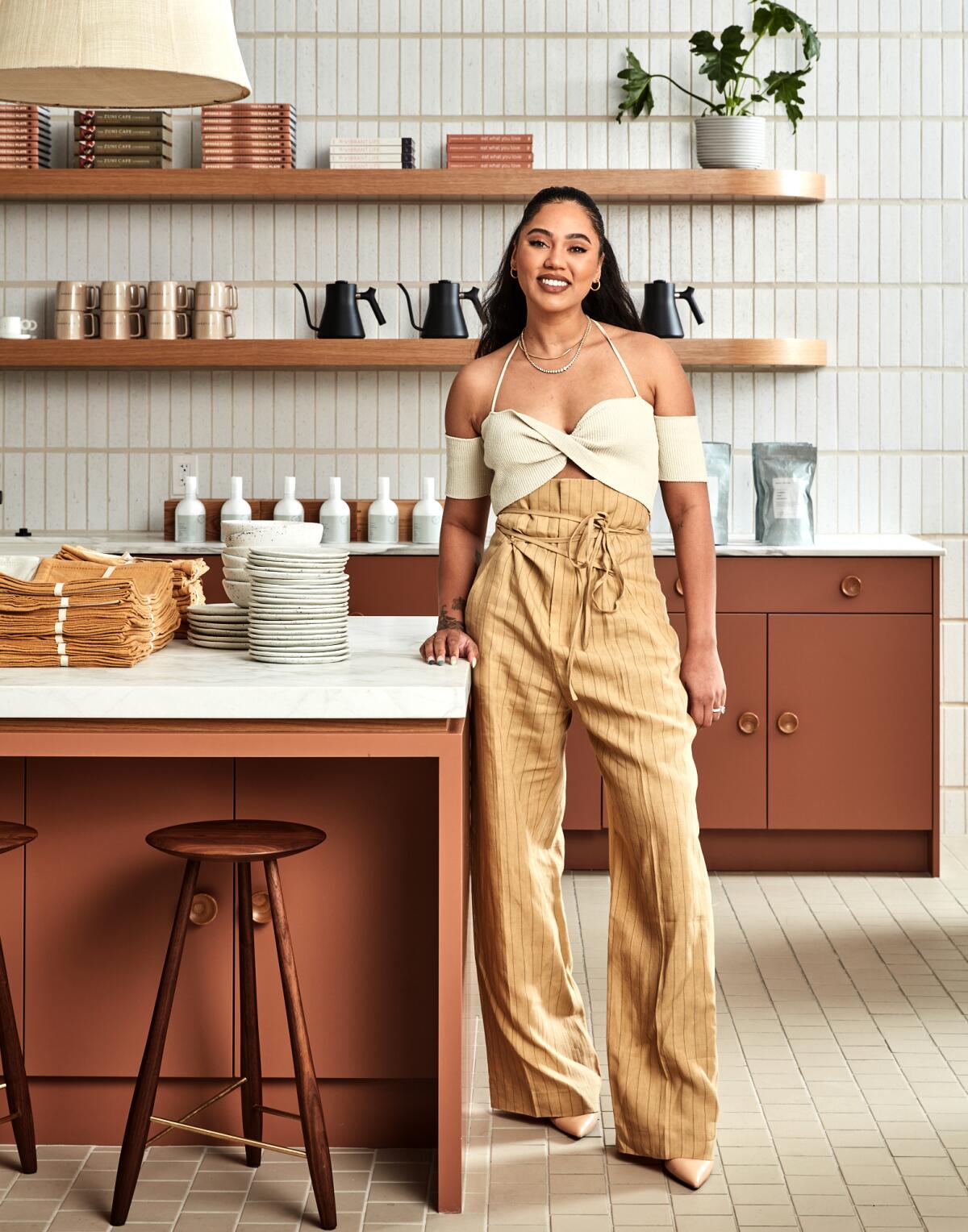 Ayesha Curry, in baggy camel pants and a white crop top, stands in a modern kitchen.