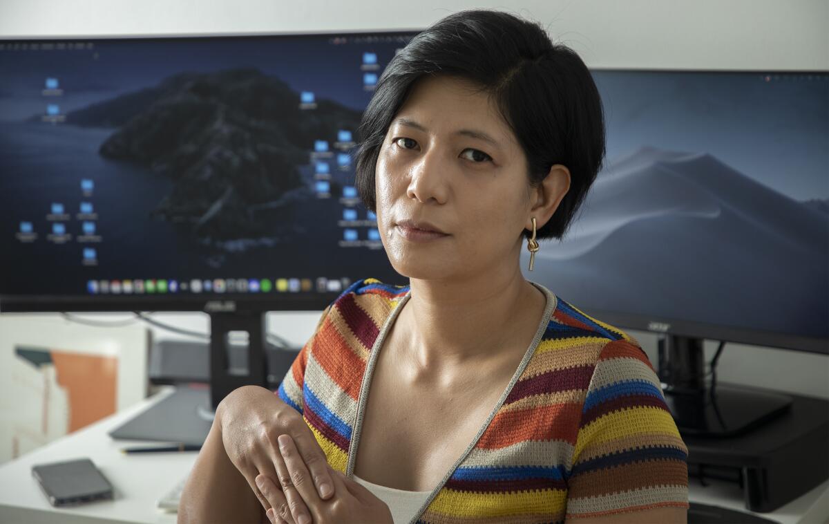 A woman in a striped shirt interlaces her fingers while sitting at a desk with two large computer monitors.