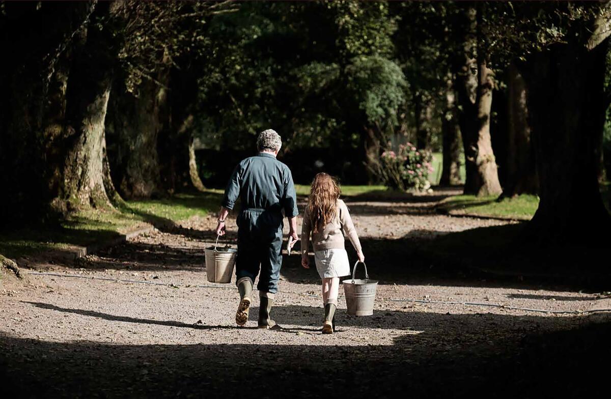 Two people, seen from behind, walk down a tree-lined path carrying buckets.