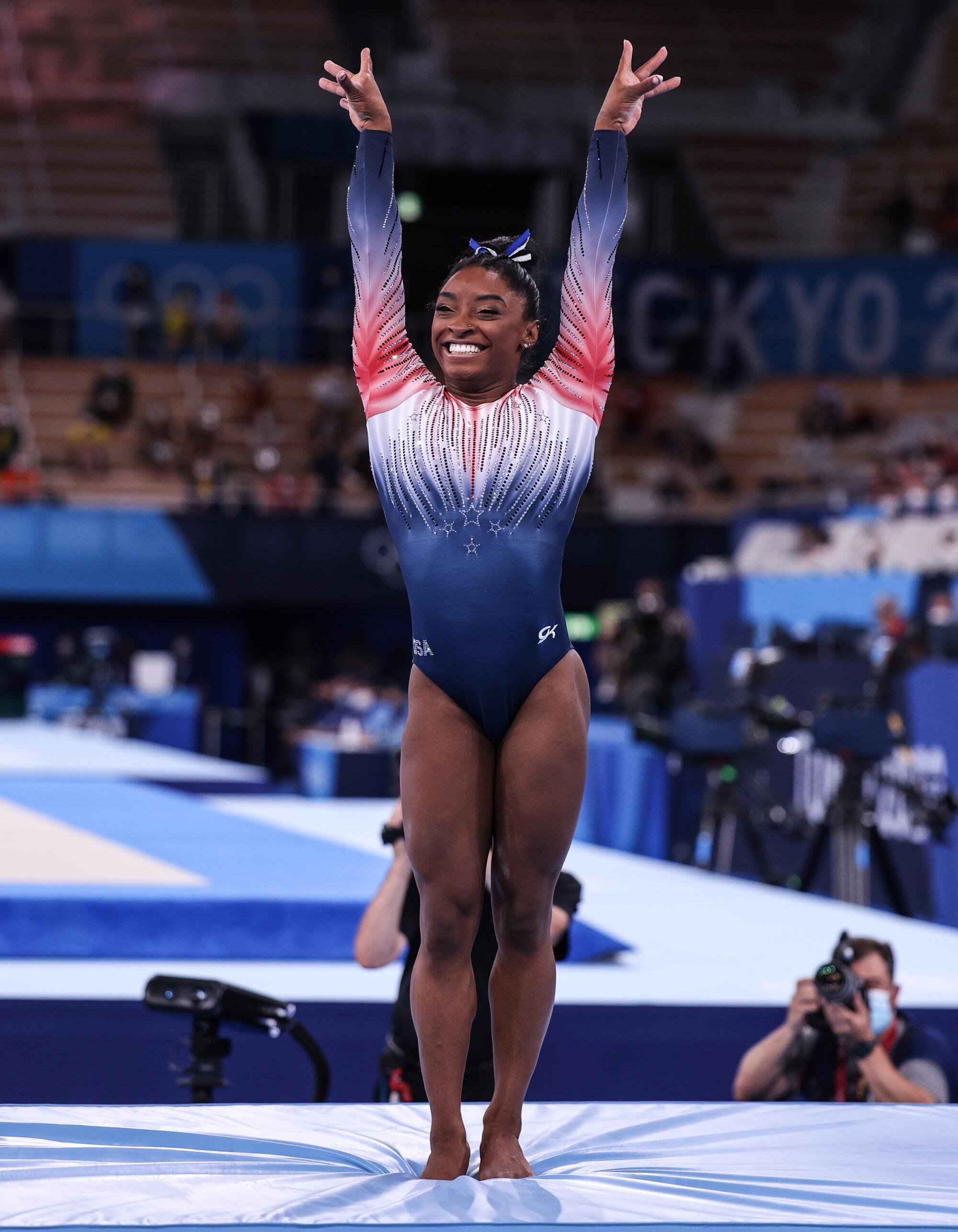 Simone Biles, smiling, stands on the mat with both arms lifted.
