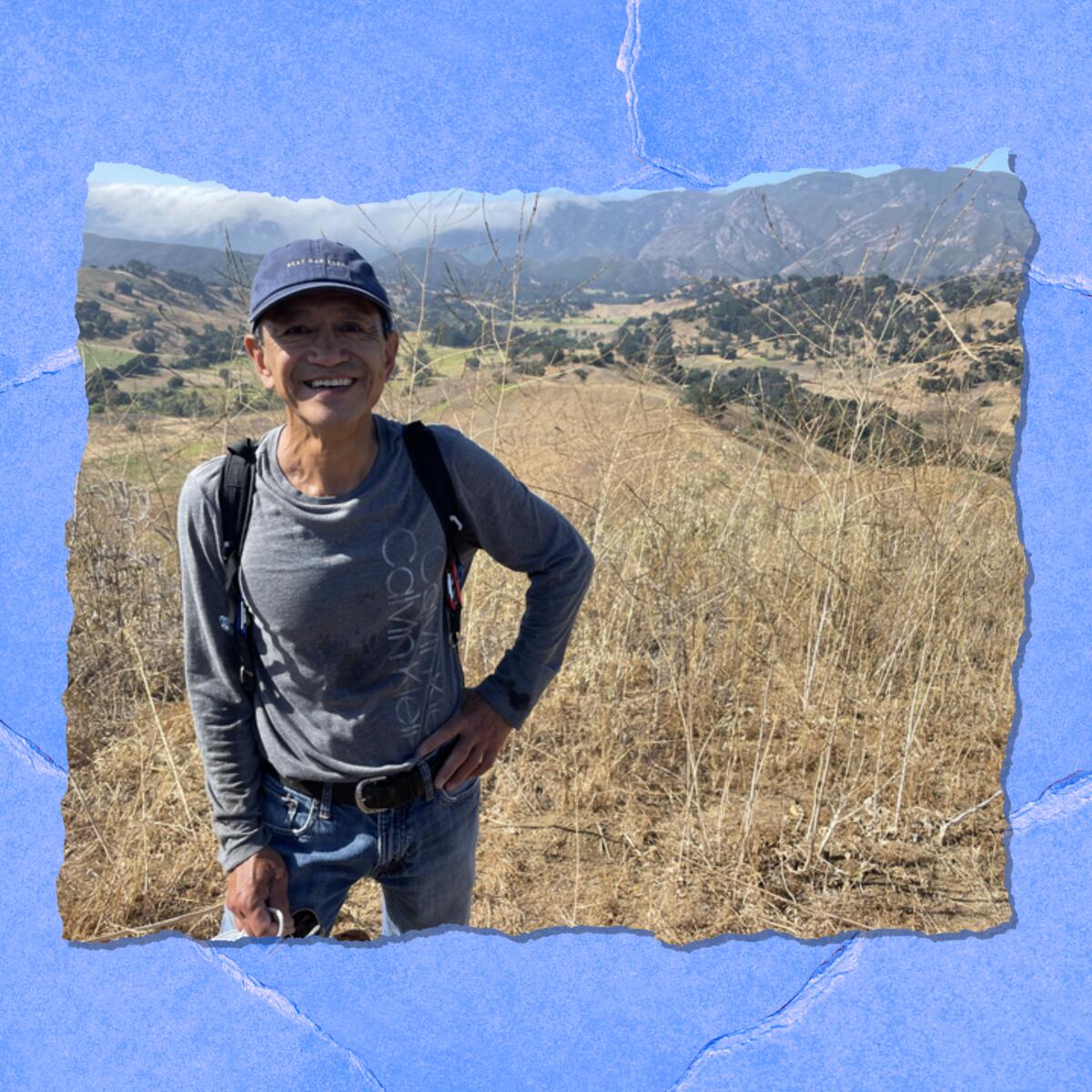 A smiling man in a ball cap and wearing a backpack stands next to scrubby land and mountains in the background.