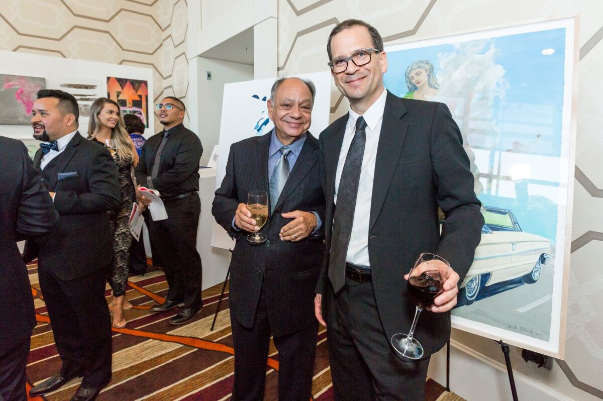 Cheech Marin and Chon Noriega smile as they stand before a work of art and hold drinks