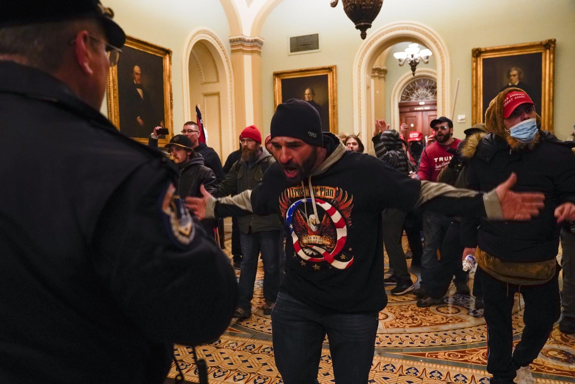 A man wearing a T-shirt referencing a conspiracy theory holds both arms wide in a gesture to a police officer.