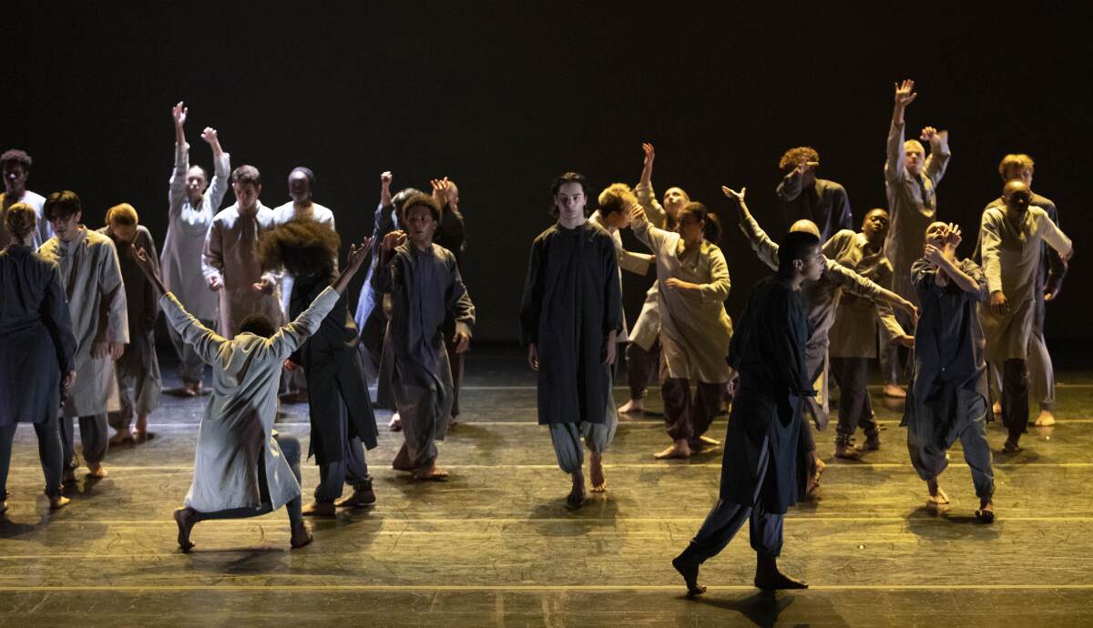 Dancers cover the stage in gray and brown clothing, some stoic and some with hands in the air.