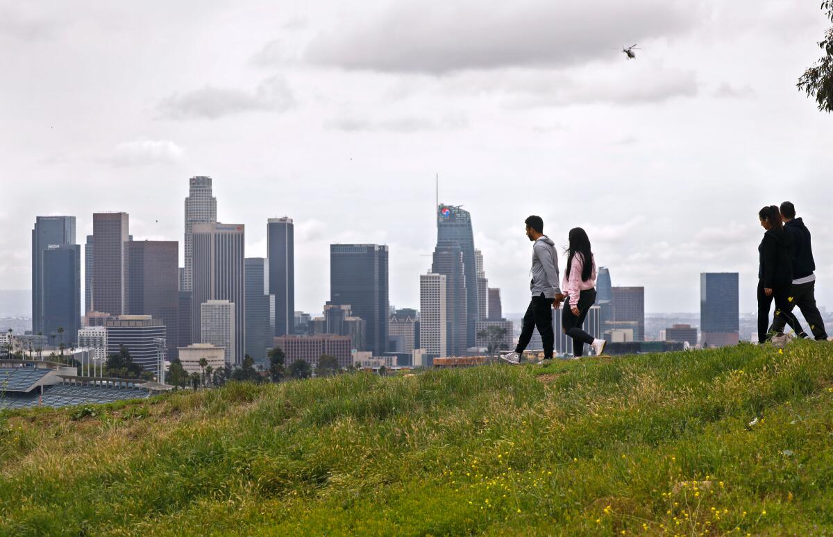 Two couples walk over a grassy area under cloudy skies with the Los Angeles skyline on the  horizon.