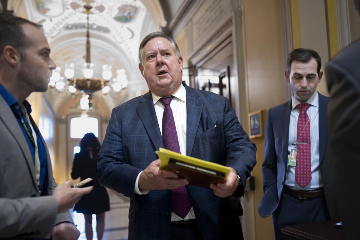 A man wearing a suit and holding a stack of papers stands in a hallway alongside two other men in suits