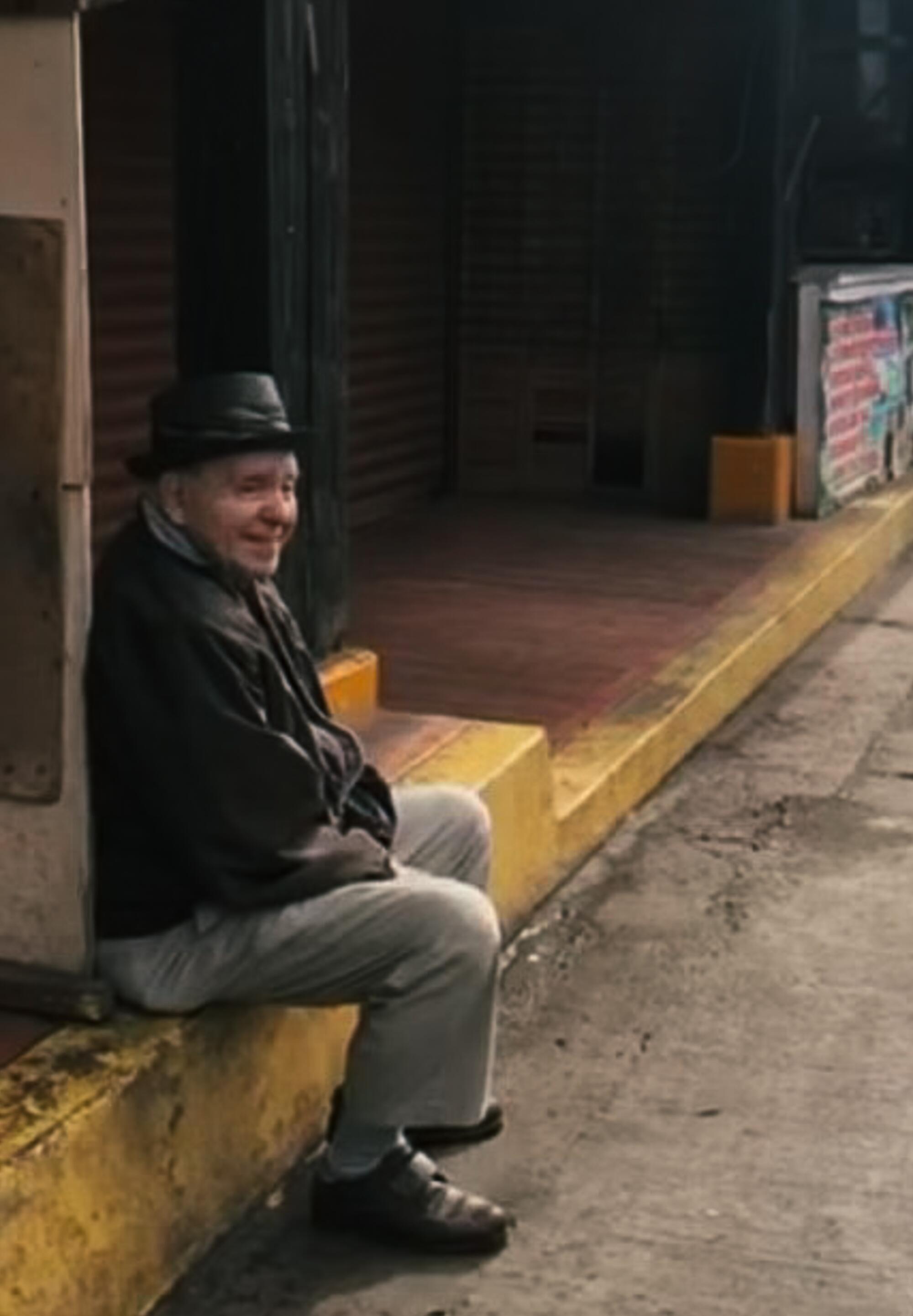 An older man sits and smiles on a curb