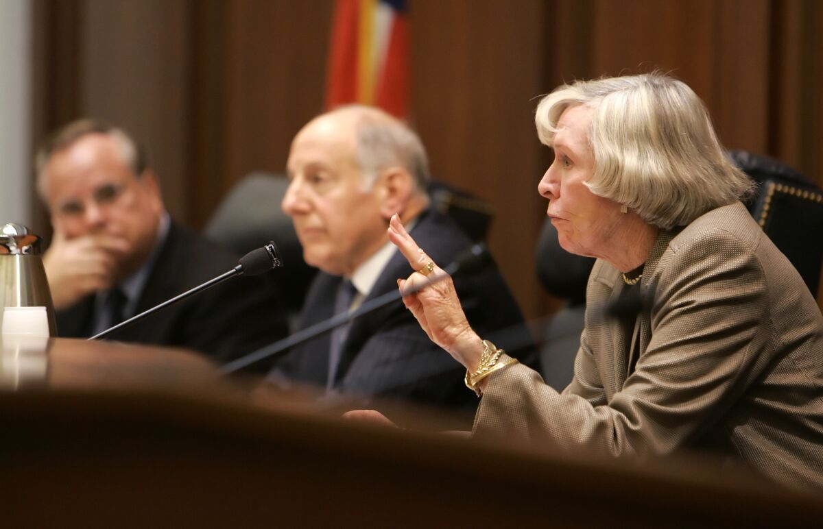 Judge Joan Dempsey Klein, with two men in the background, speaks into a microphone.