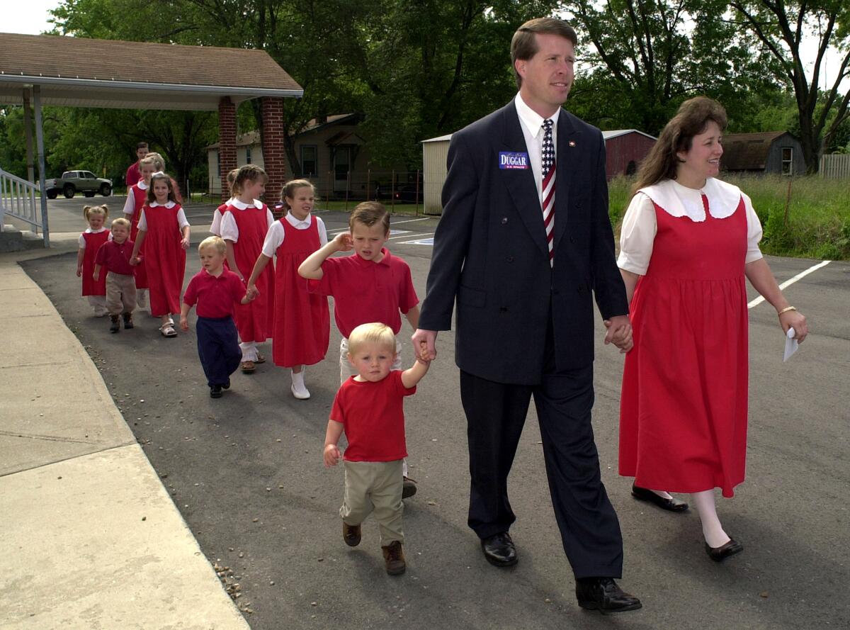 A man in a suit and a woman in a red dress lead their 12 children across the street.