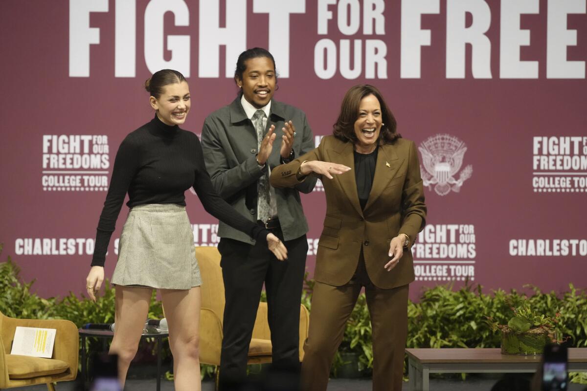 Vice President Harris laughs onstage, joined by a young woman and man, with a "Fight for Our Freedoms College Tour" backdrop