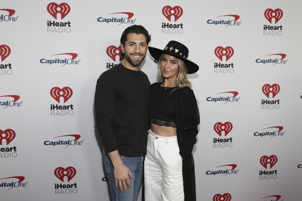 Jason Tartick in a black sweater and jeans next to Kaitlyn Bristowe in a black wide-brimmed hat and sweater and white jeans