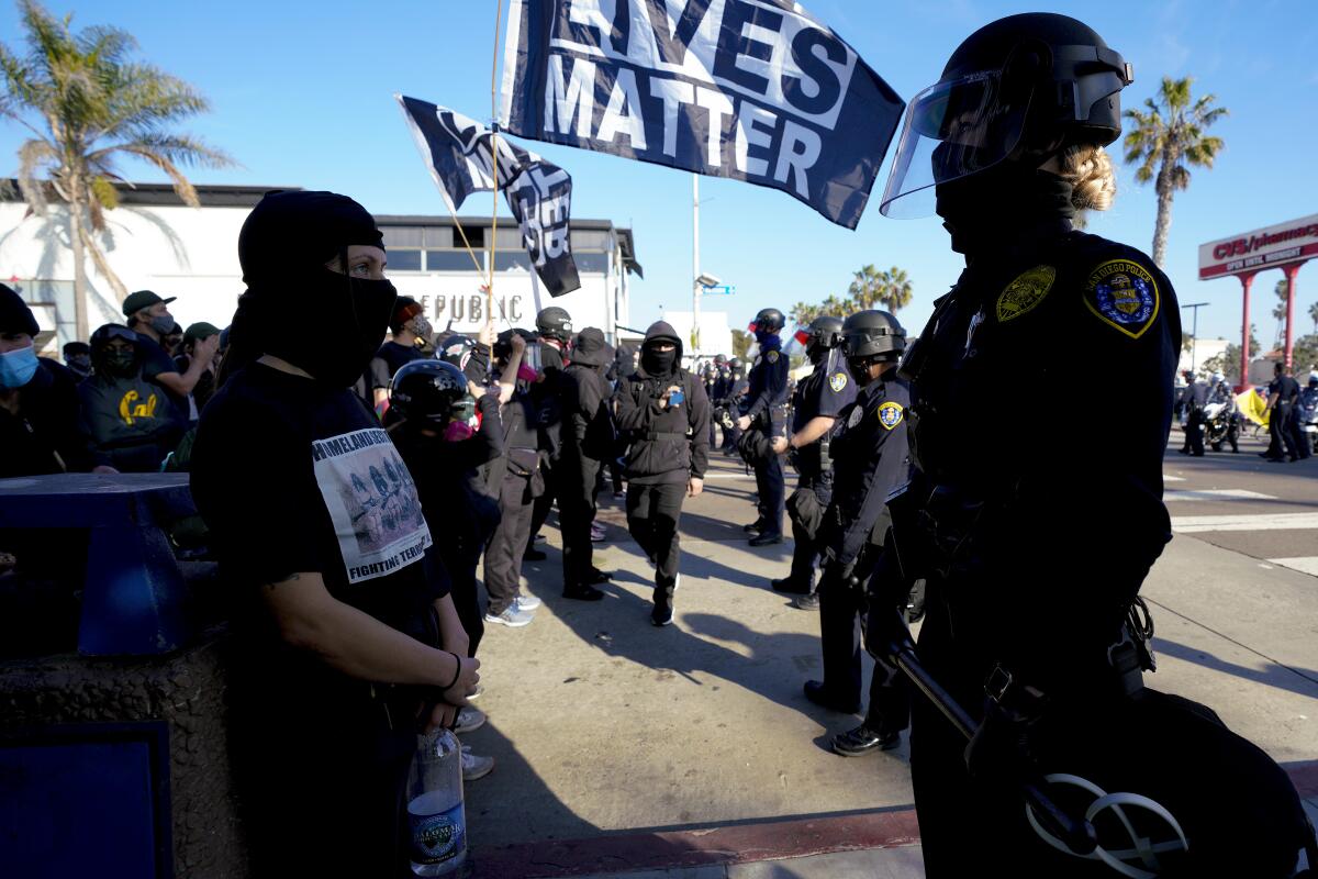 Police officers in riot gear face a group of people, most of them wearing black and covering their faces with masks.