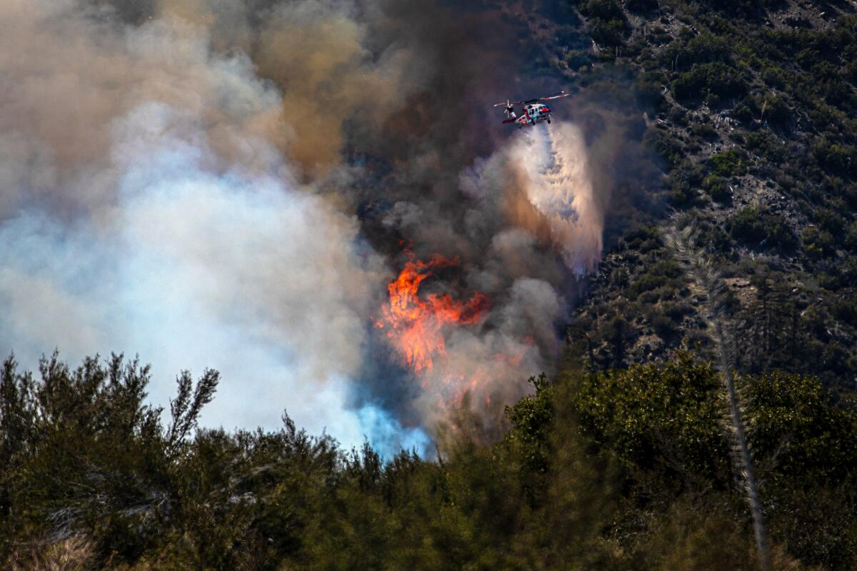 Flames and smoke rise from a wildfire in mountainous forest land as a helicopter drops water on the site