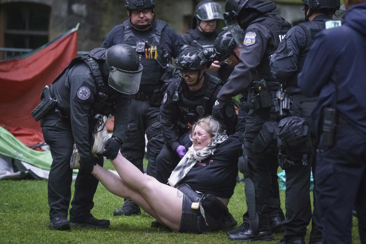 A group of police in helmets and body armor hold a young woman's legs and arms as they lift her from the ground. 