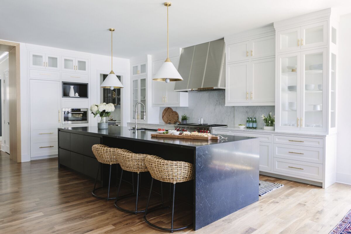 This image released by Fresh Twist Studio shows a kitchen design with woven Palecek kitchen stools and a striking midnight blue stone island to balance the extensive Shaker-style white and glass cabinetry. The brass-toned lining of the pendant fixtures from Circa Lighting is echoed in the cabinet hardware. (Aimee Mazzenga/Fresh Twist Studio via AP)