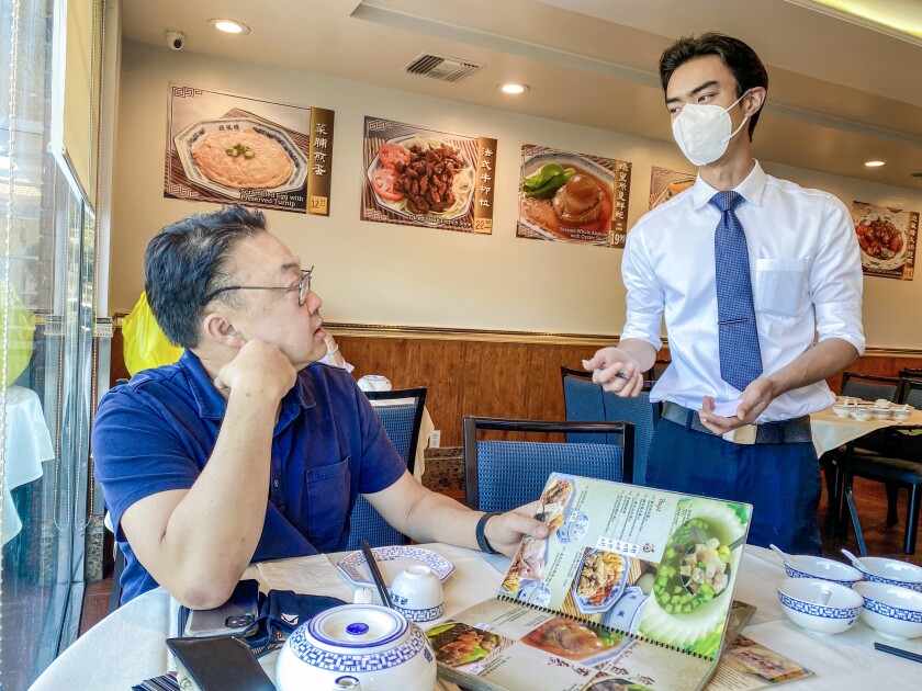 A man sitting at a round dining table holds a menu and looks at a waiter standing nearby.