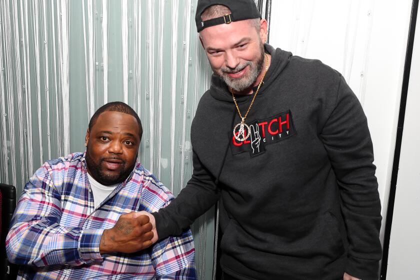 Big Pokey sits and shakes hands with Paul Wall, who is standing.