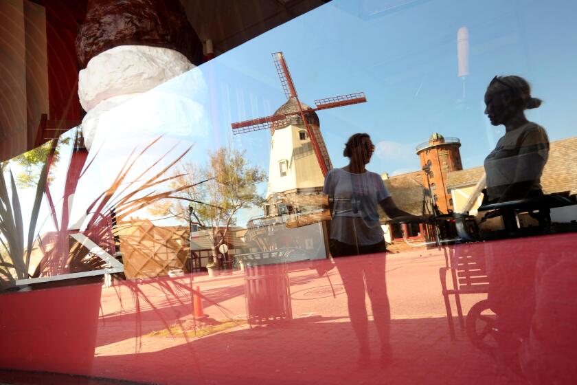 SOLVANG, CA - APRIL 16, 2020 - - Locals Alicja Clarke, right, and Jusyna Zimkowski, enjoy a conversation while reflected in a nearby storefront window along with the iconic windmill on Alisal Road in Solvang, California on April 16, 2020. Clark has not been working since The Copenhagen House had to close due to the coronavirus pandemic. Solvang is a tiny town of 5,300 people nestled in the Santa Ynez Valley wine country, about a half-hour north of Santa Barbara. Tourists - mainly those who make the short drive from Los Angeles and Orange counties -- are the engine of an economy than generates nearly $200 million in economic activity a year. And without it hundreds in Solvang and the surrounding bedroom communities have lost their jobs while the city is losing $500,000 in tax revenue a month. (Genaro Molina / Los Angeles Times)