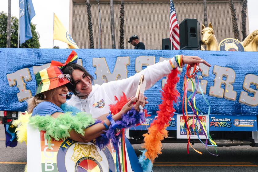 Two women dressed in rainbow outfits and accessories taking a selfie in front of a parade float on the street.