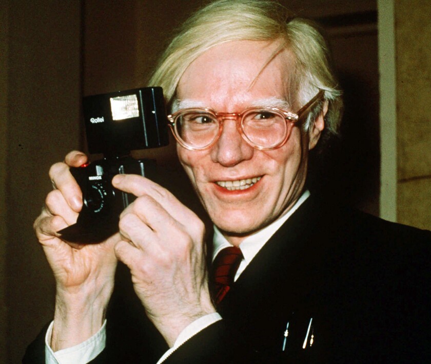 Pop artist Andy Warhol smiles for the camera while holding another camera in 1976.