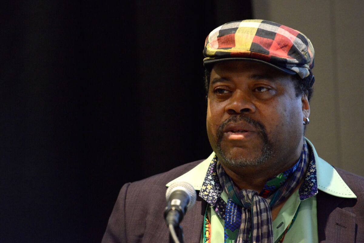 Greg Tate is seen wearing a plaid newsboy cap and a printed scarf while standing before a microphone.