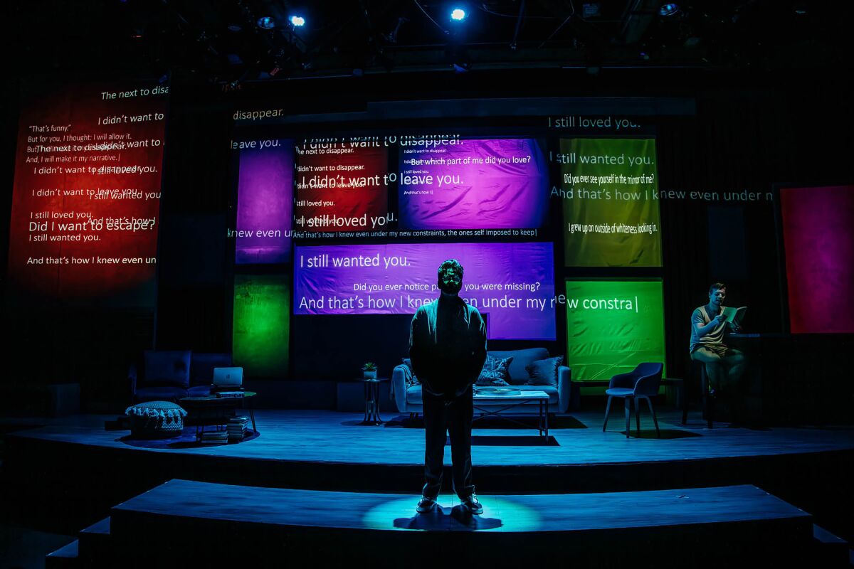 man stands on a stage alone as words are projected on colorful screens behind him