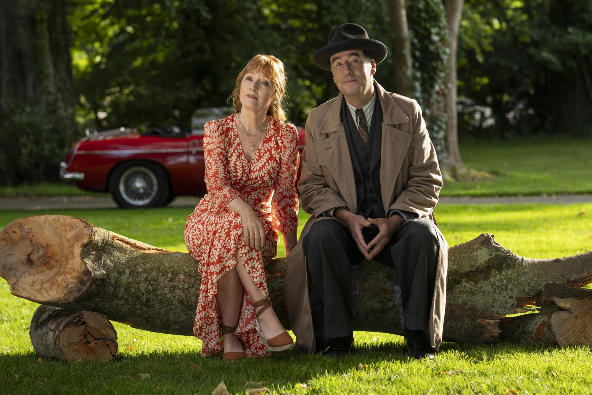 A woman in a red floral dress sits next to a man in a suit and overcoat on a felled tree.