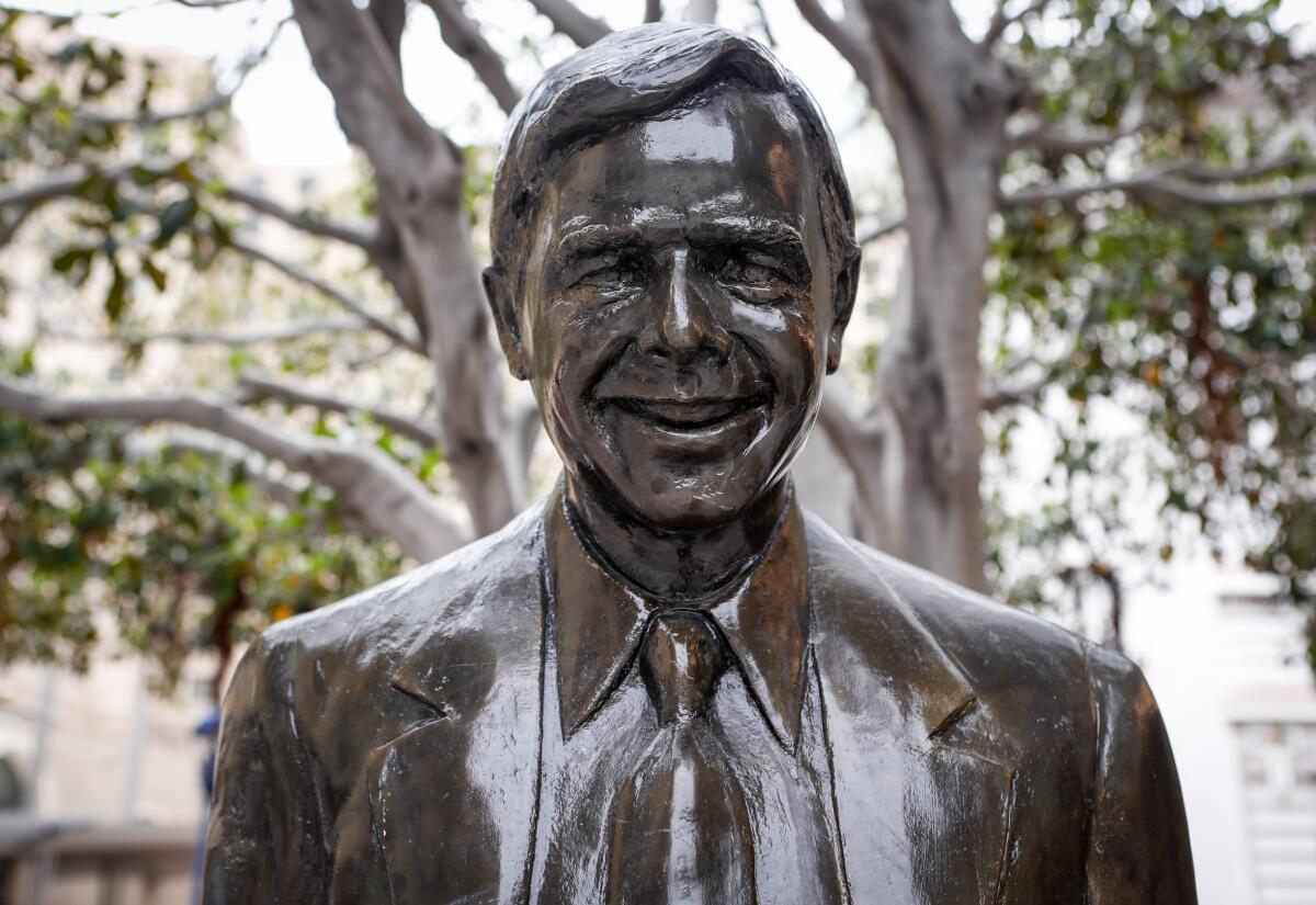 A bronze statue of a smiling man in a suit is seen from the shoulders up in front of a leafy tree.