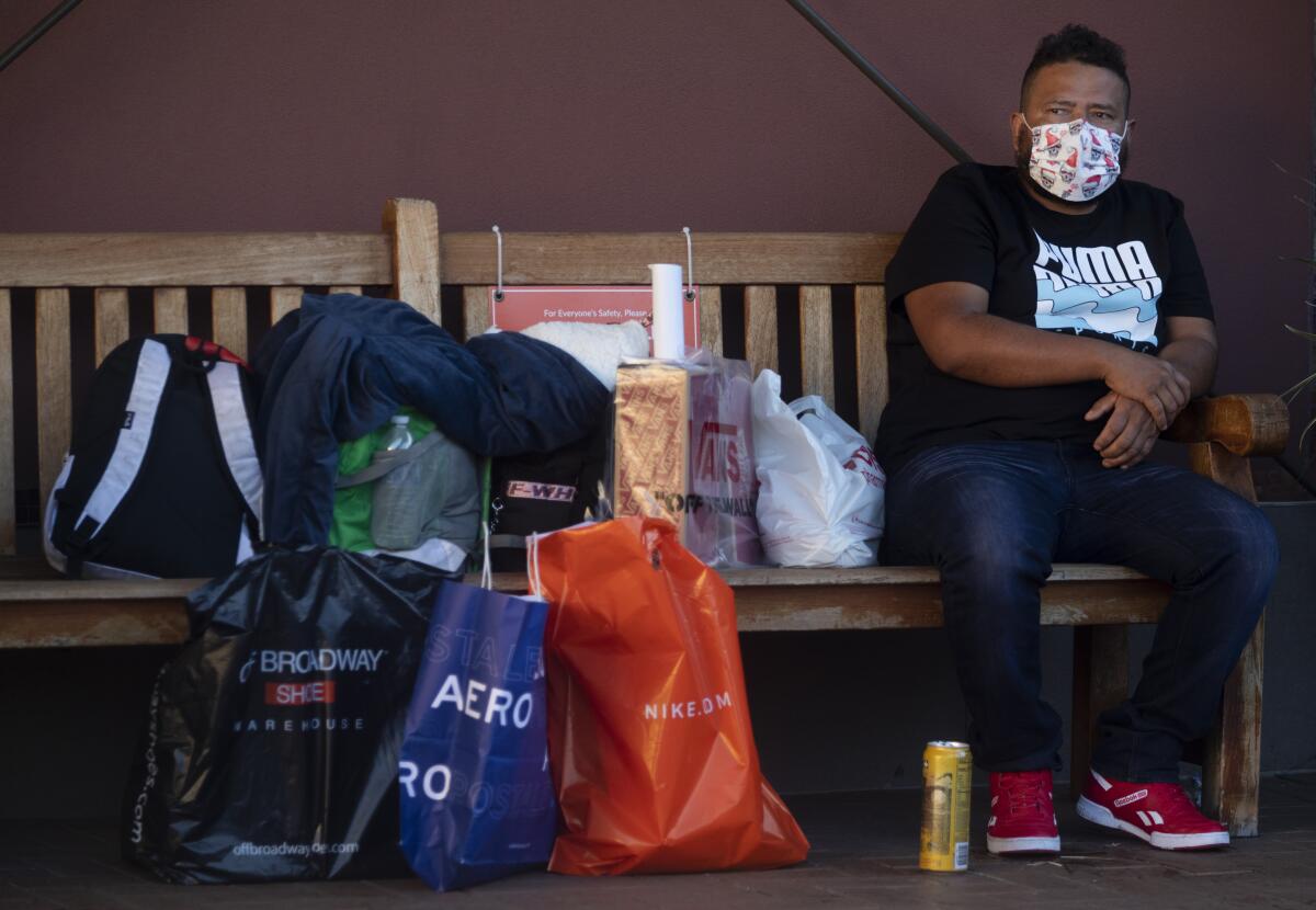 A man in a mask sits on a bench with a pile of shopping bags and backpacks next to him