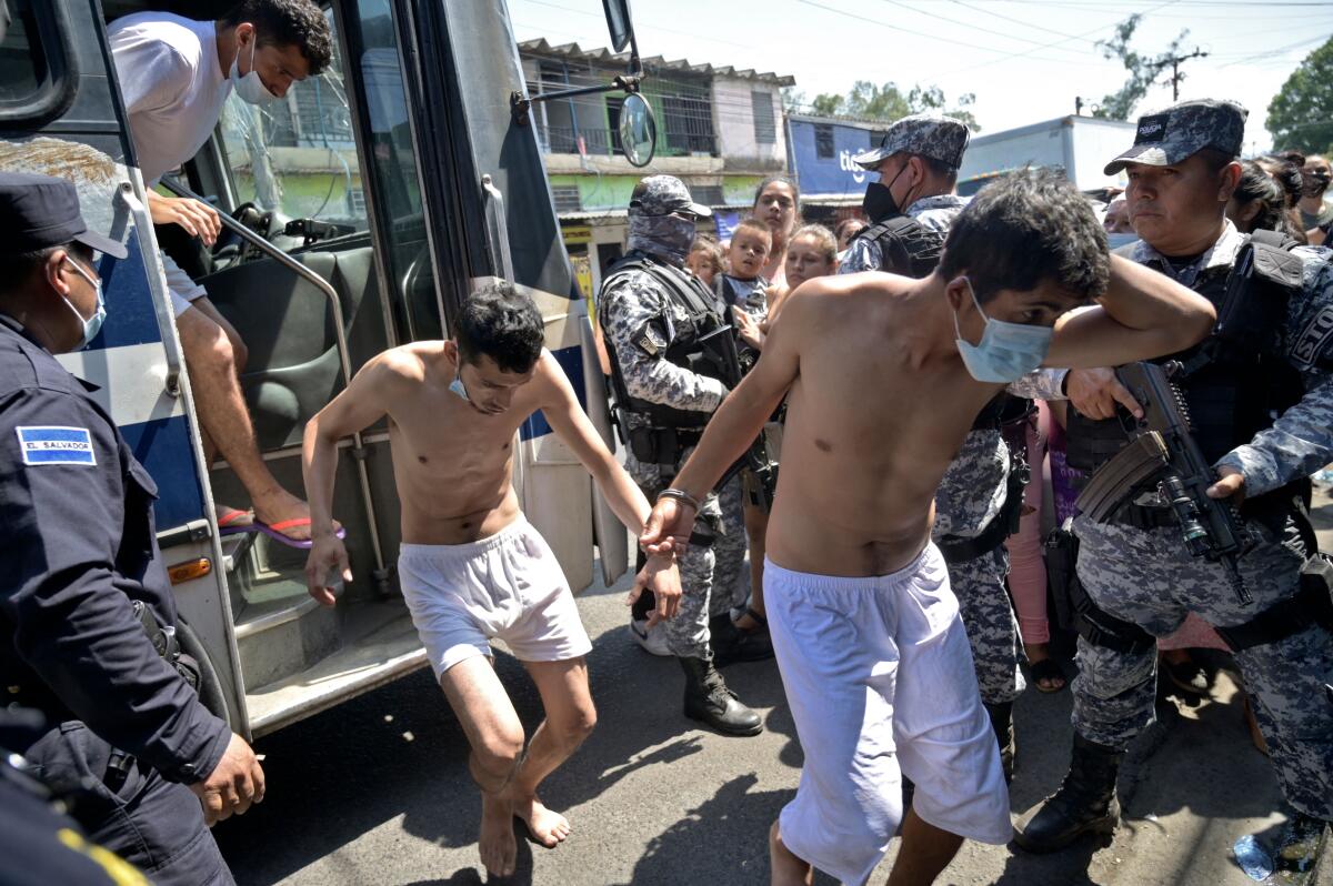 Men in white shorts and no shirt disembark from a bus with armed men in fatigues standing watch 