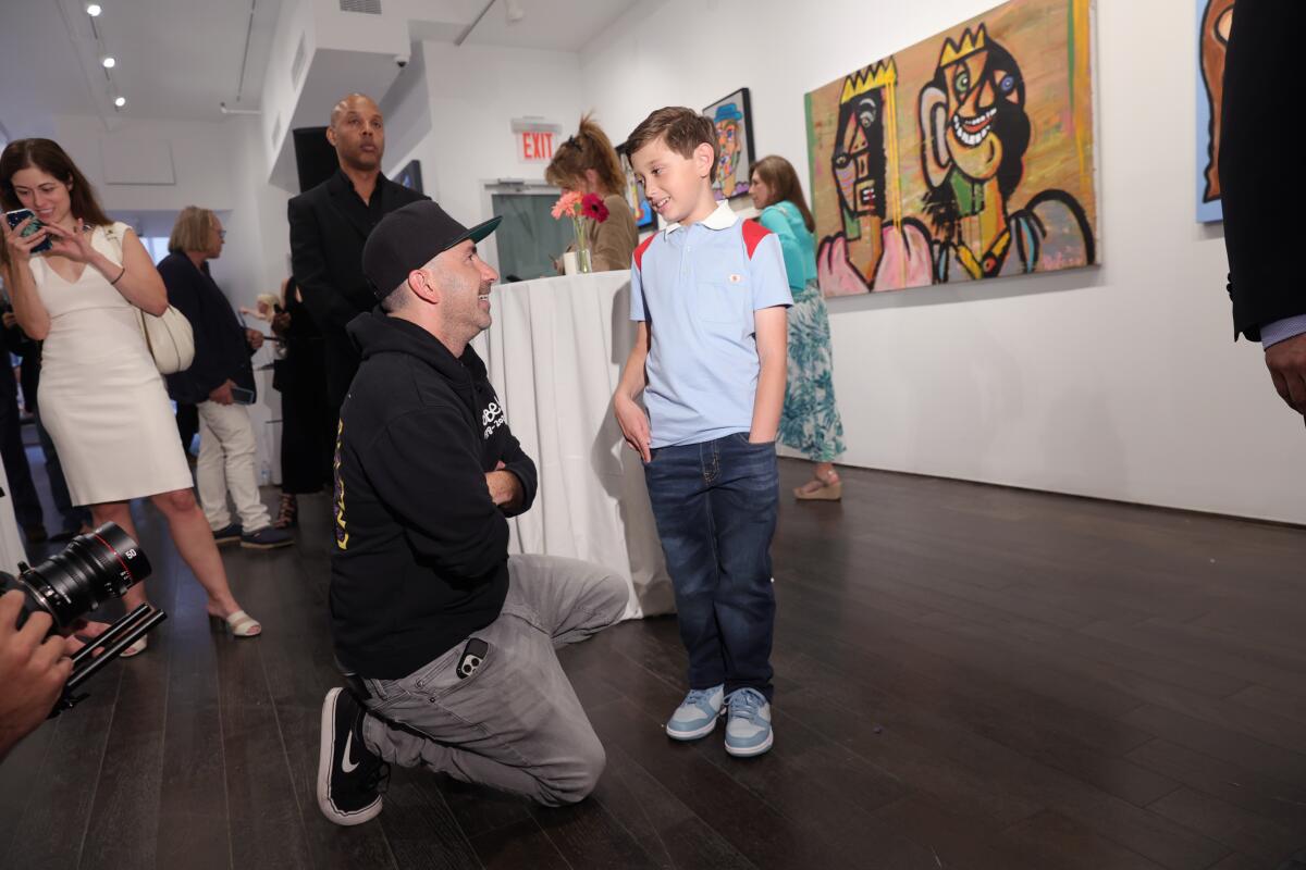 A man in a ballcap kneels to talk to a young artist, a boy wearing a polo shirt and jeans, with art in the background