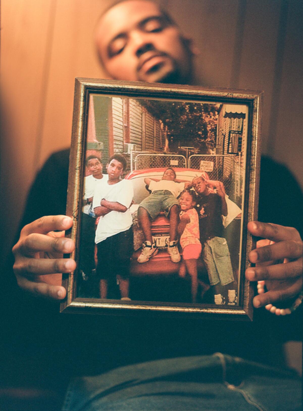 A man holds a framed photo of his family leaning on the back of a parked car