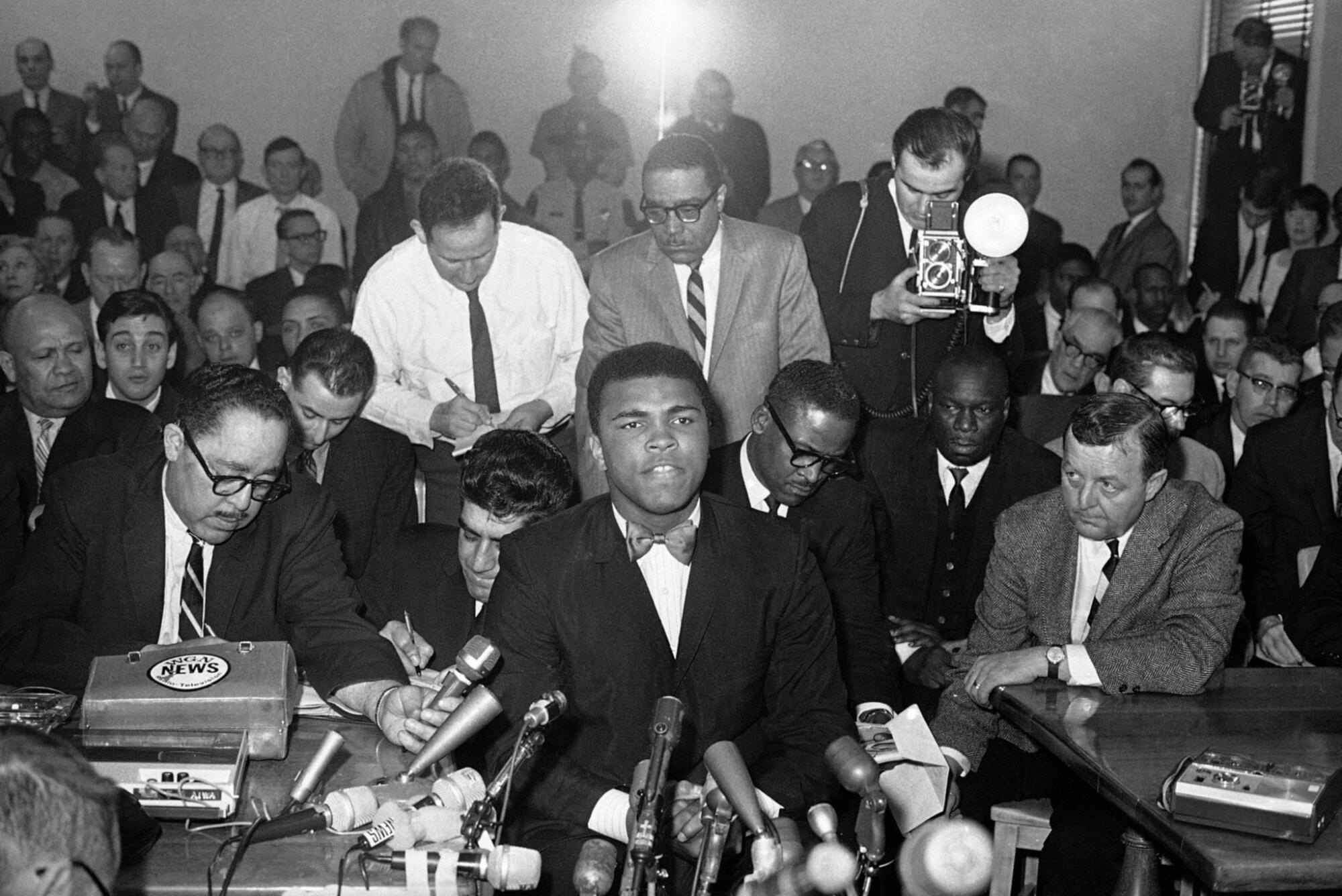 A black-and-white photo of Muhammad Ali in a dark suit sitting at several microphones, surrounded by members of the media