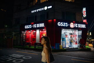 A woman walks by a building with a sign that reads GS25 x FC Seoul at night.