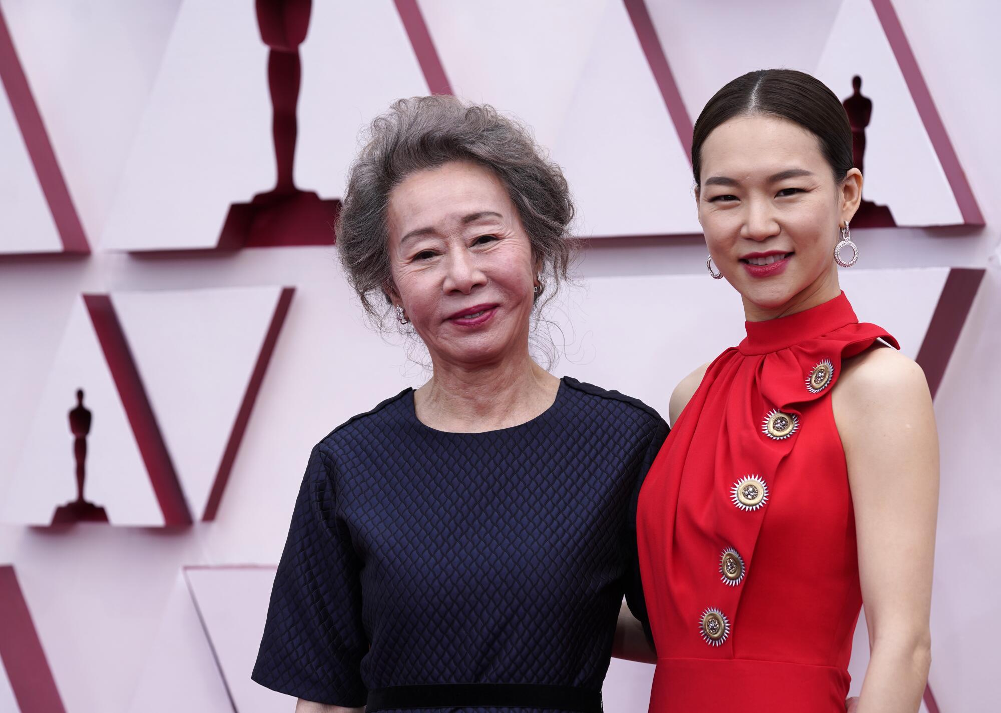 Yuh-Jung Youn wears a dark dress, and Han Ye-ri wears a high-neck red dress with large buttons