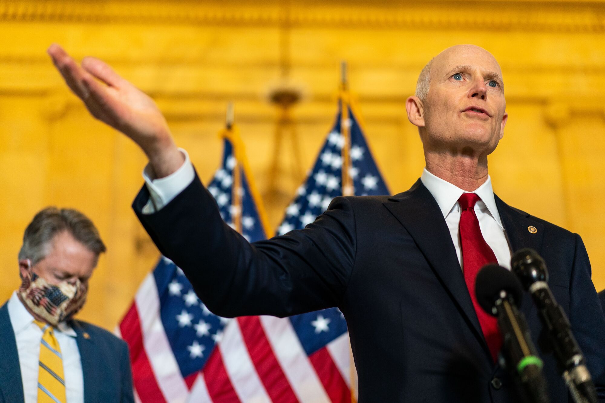 Rick Scott in a suit and red tie speaking into microphones while gesturing with his right arm raised.