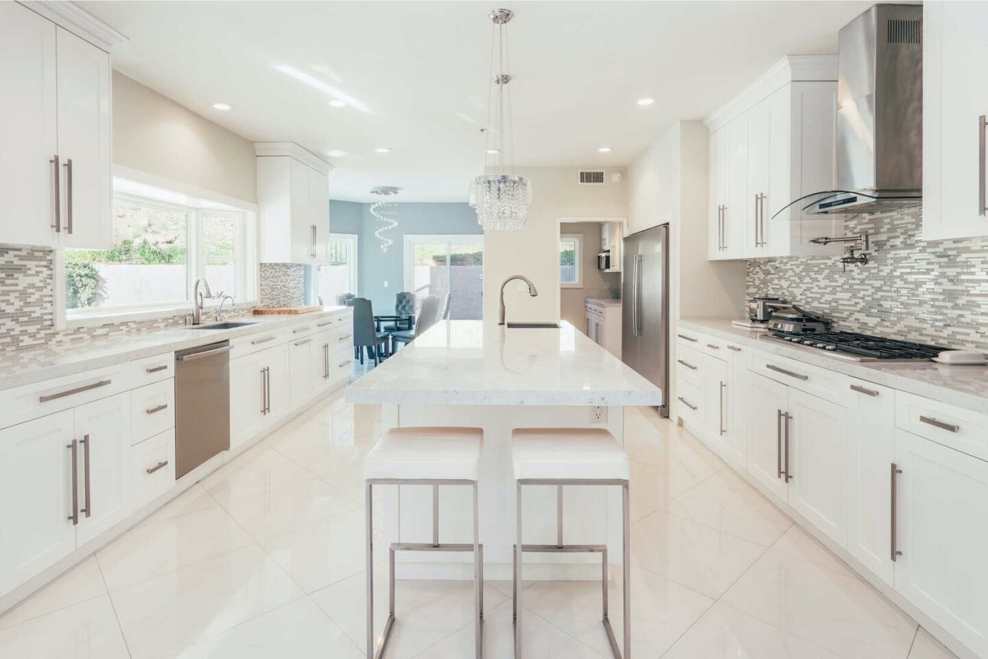 A long kitchen with long, marble-topped center island, white cabinets and tile backsplashes.