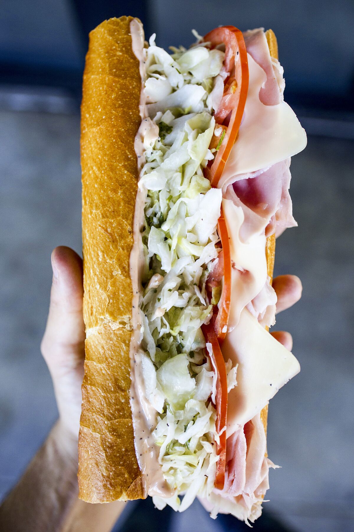 A photo of a hand holding a large ham and cheese sandwich on baguette, the Heavenly Ham N Swiss.