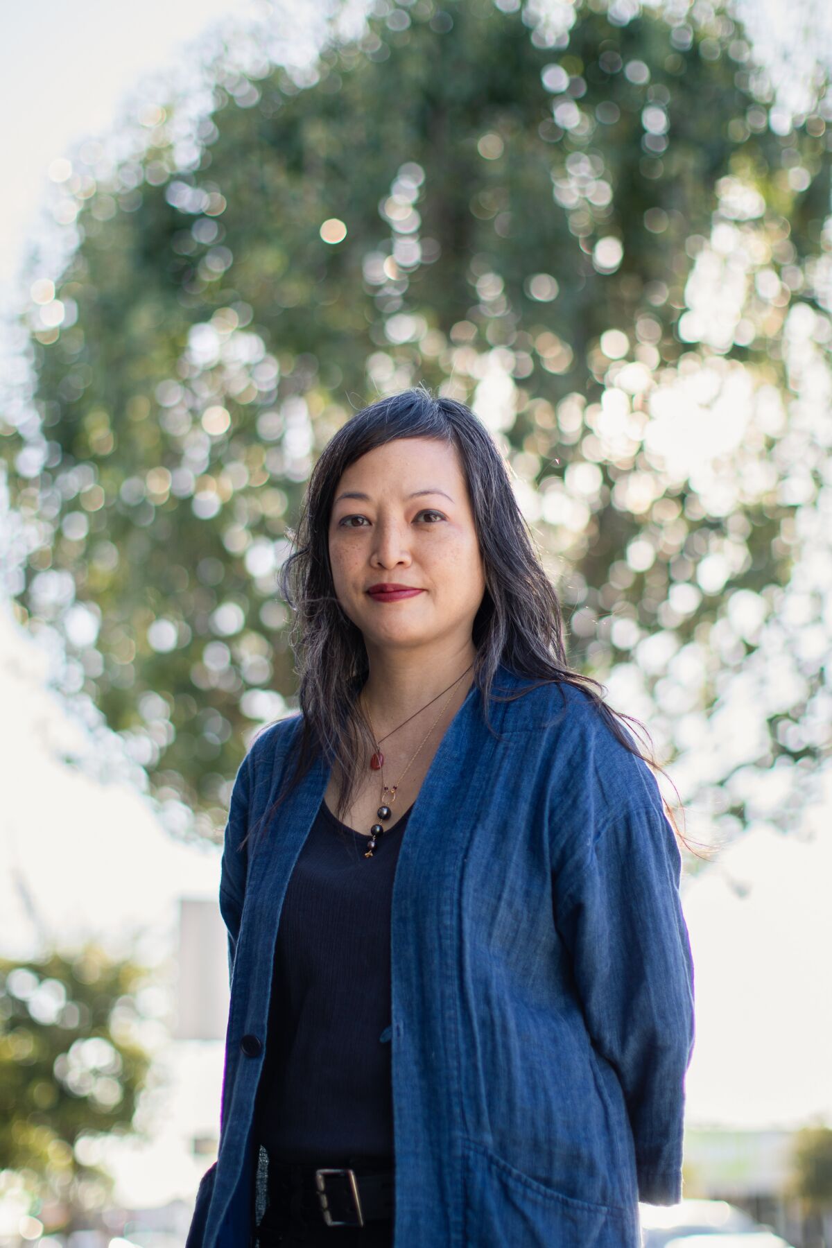 Carla Ching, in a blue shirt, is seen standing outdoors, framed by the branches of a tree.