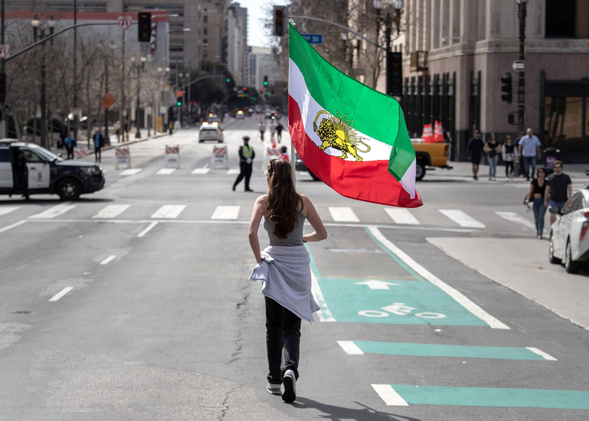 A woman carrying a large Iranian flag walks down the middle of a street.