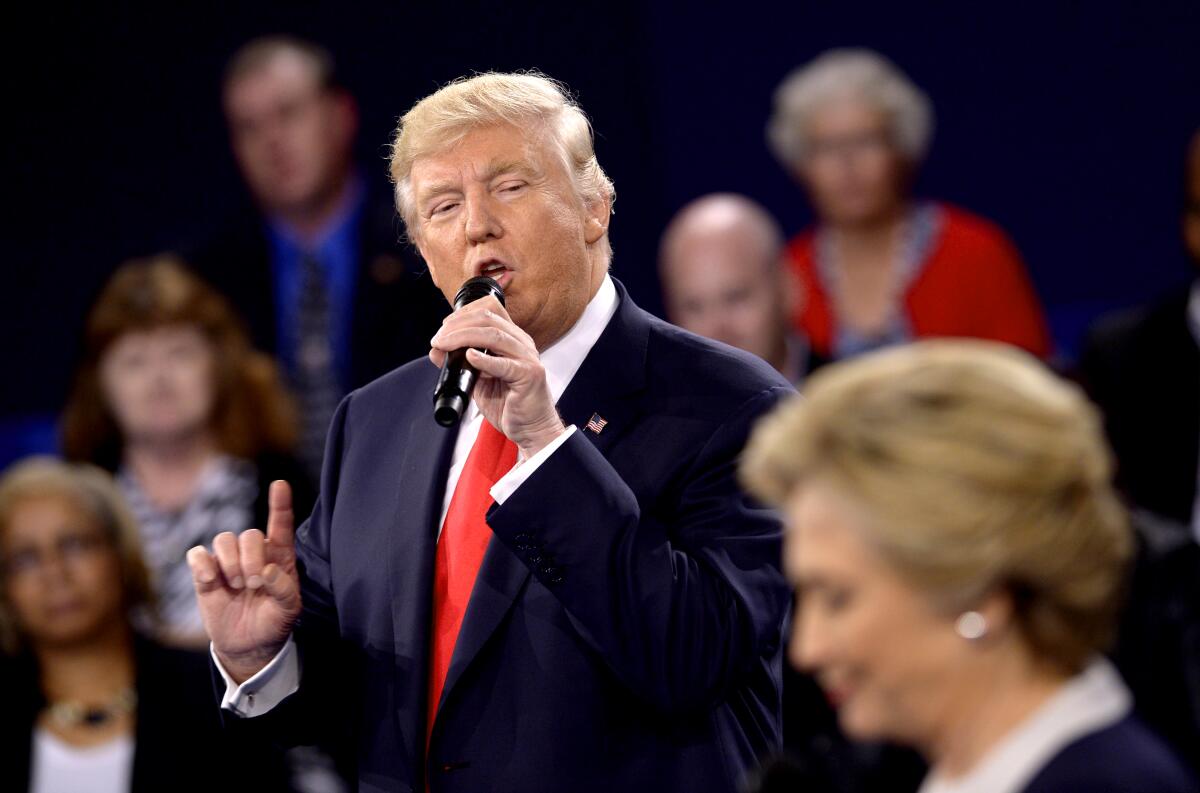 Donald Trump holds up a finger, looking at Hillary Clinton as he speaks into a microphone, audience members in the background