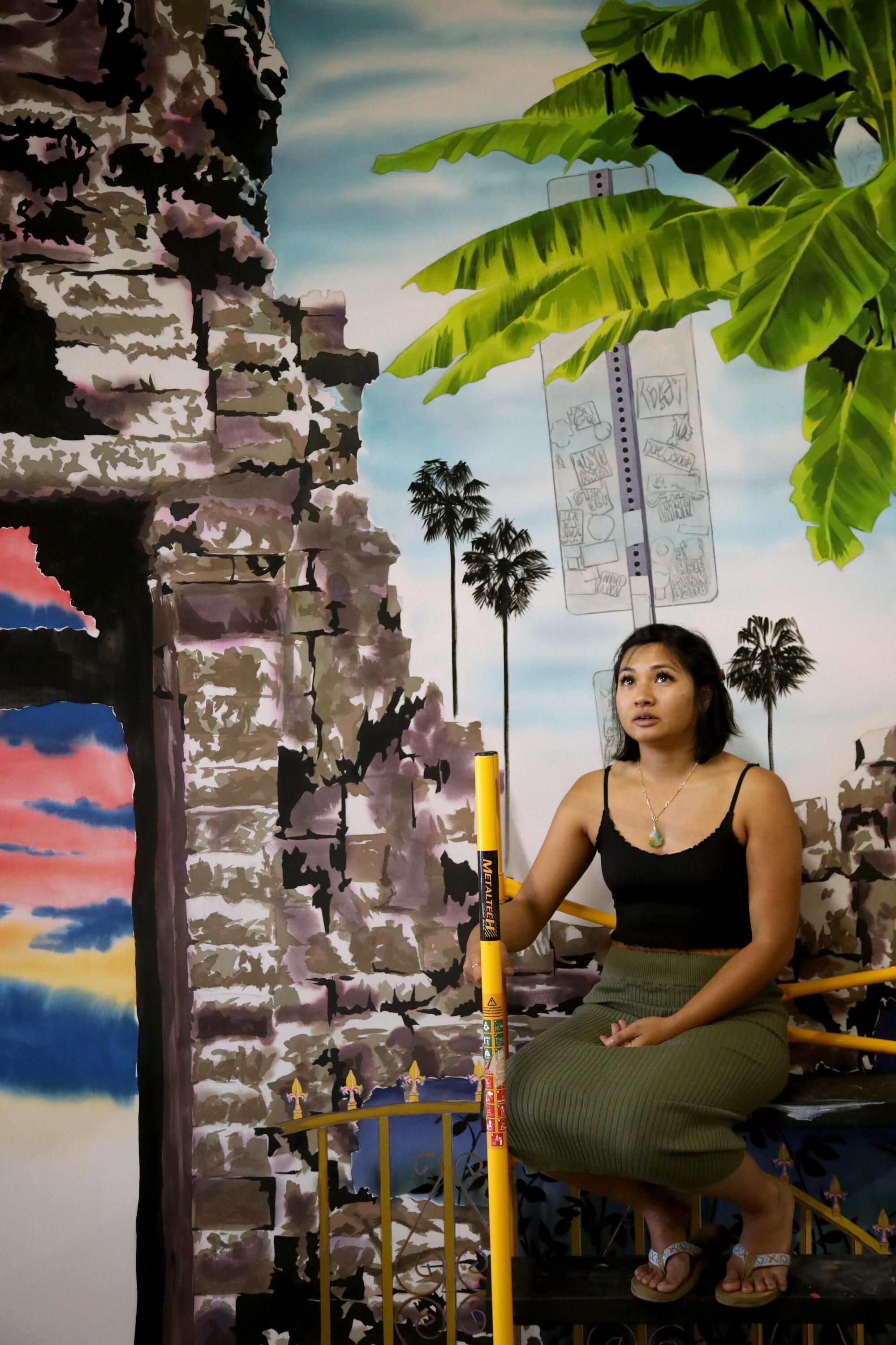 Tidawhitney Lek, a Cambodian American woman, sits on a scaffold in a studio lined with paintings.