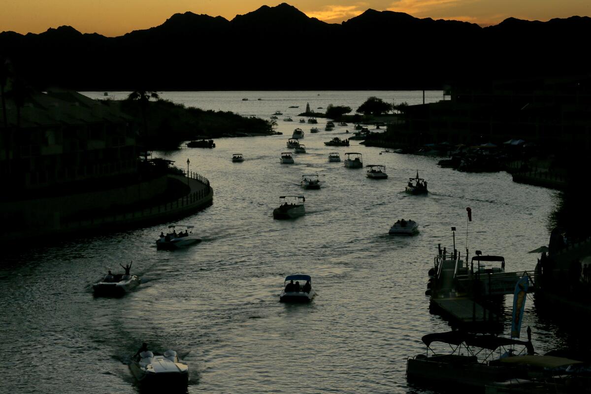 More than a dozen boats winding through a curving channel in dim light as the sun sets behind mountains in the distance