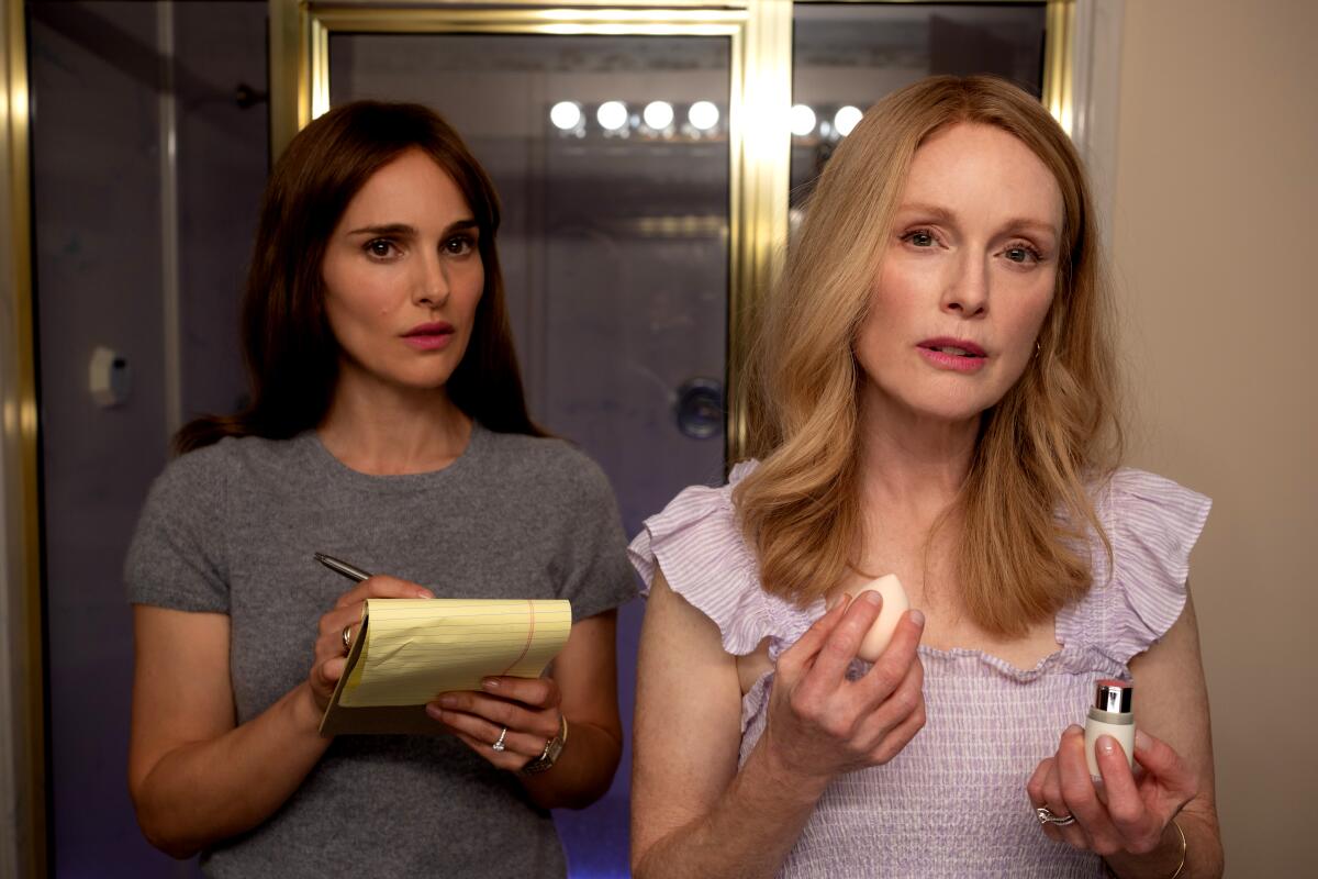 A woman holding a notepad watches an older woman putting on her makeup in a scene from "May December."