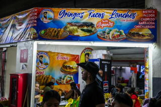 A man in dark clothes and a hat near an eatery with banners depicting various dishes