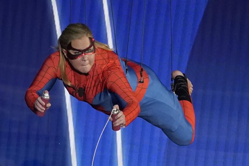 A woman with blond hair wearing a Spider-Man costume while suspended from a wire