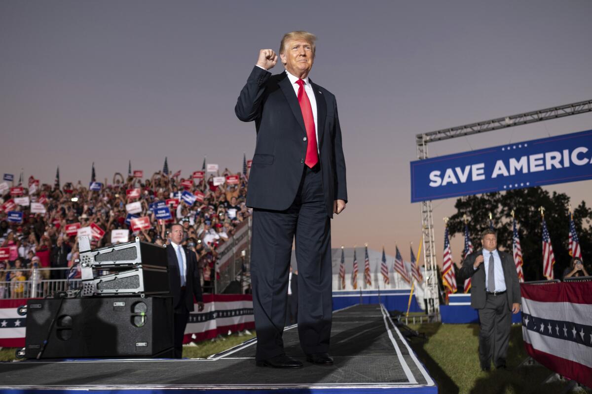 A man in a dark suit and red tie with a clenched fist leading a rally in front of a sign that says “Save America.”